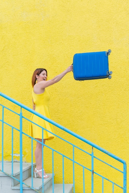 Uma mulher ruiva feliz em um vestido amarelo está descendo as escadas e balançando alegremente uma mala azul A garota está se preparando para a viagem Conceito de férias de verão