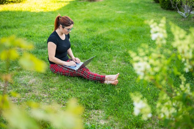 Uma mulher ruiva senta-se na grama verde de um parque e faz compras online A garota mantém distância social e trabalha ao ar livre em um laptop Um aluno está estudando remotamente em um computador