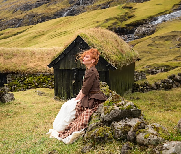 Uma mulher ruiva sentada em uma cerca de pedra com roupas antiquadas. Aldeia de Saksun, Streymoy, Ilhas Faroe