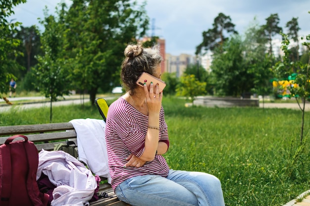 Foto uma mulher se senta em um banco de parque e fala em um telefone celular, encostando-o no ouvido, copie o espaço.