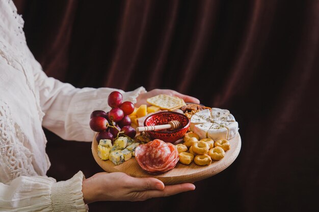 Uma mulher segura nas mãos uma tábua de madeira redonda com aperitivos de vinho cortando salsicha de queijo e frutas