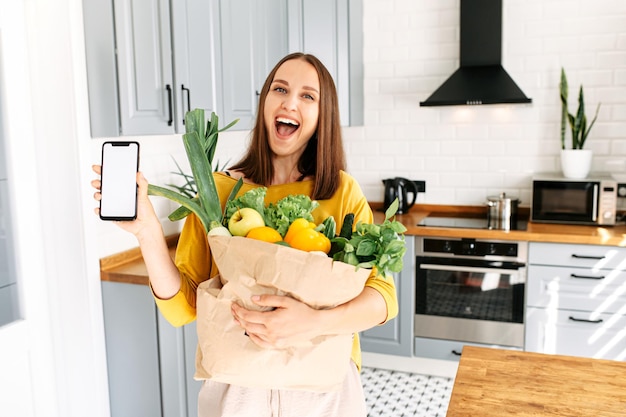 Uma mulher segura um telefone e um saco cheio de vegetais