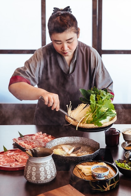 Uma mulher segurando legumes em panela quente por pinças com Wagyu A5 carne e Kurobuta fatiado em Shabu.