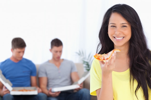 Uma mulher segurando pizza na mão enquanto seus amigos se sentam atrás dela