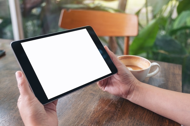 uma mulher segurando preto tablet pc com espaço em branco com uma xícara de café na mesa de madeira