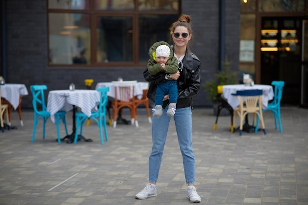 Uma mulher segurando um bebê em uma jaqueta de couro preta e jeans azul na frente de um restaurante