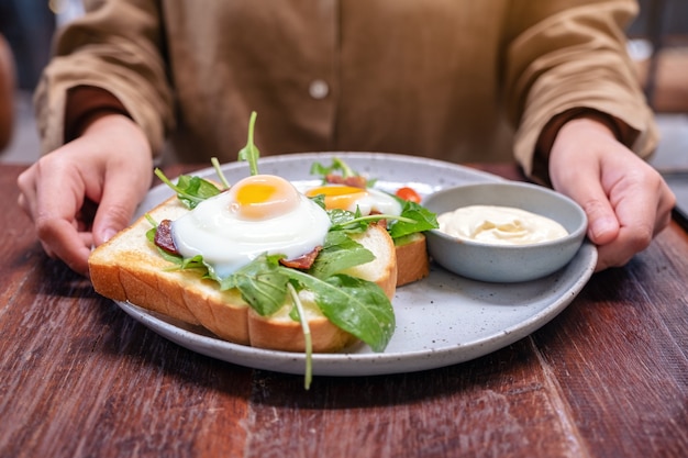 Uma mulher segurando um prato de sanduíche de café da manhã com ovos, bacon e creme de leite na mesa de madeira