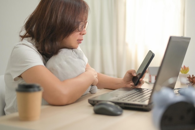 Uma mulher segurando um smartphone e usando um laptop para trabalhar em casa, vestindo uma camisa branca com óculos e abraçando um travesseiro, uma xícara de café e um laptop em cima da mesa, escritório em casa.