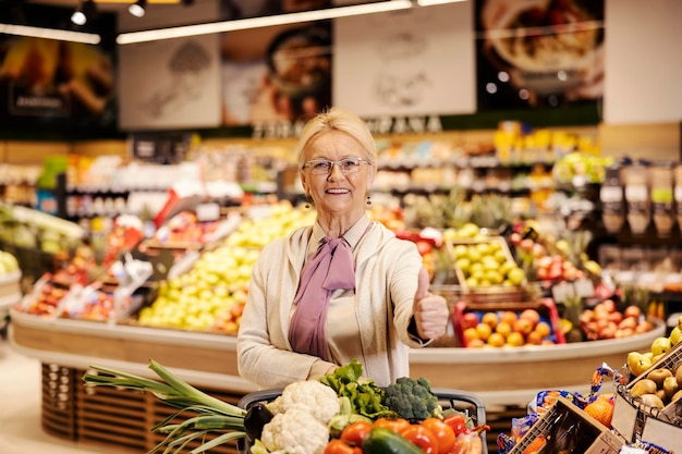 Uma mulher sênior está comprando no supermercado e dando polegares para cima enquanto sorri para a câmera