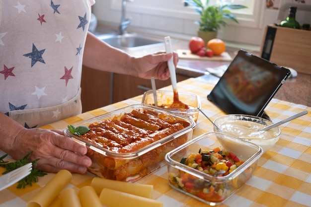 Uma mulher sênior preparando uma panela cheia de canelone artesanal recheado com legumes e ragu Seguindo a receita italiana no tablet