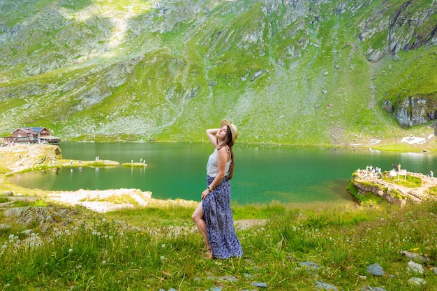 Uma mulher sensual em um vestido longo e chapéu de palha fica às margens do lago balea nas montanhas romenas.