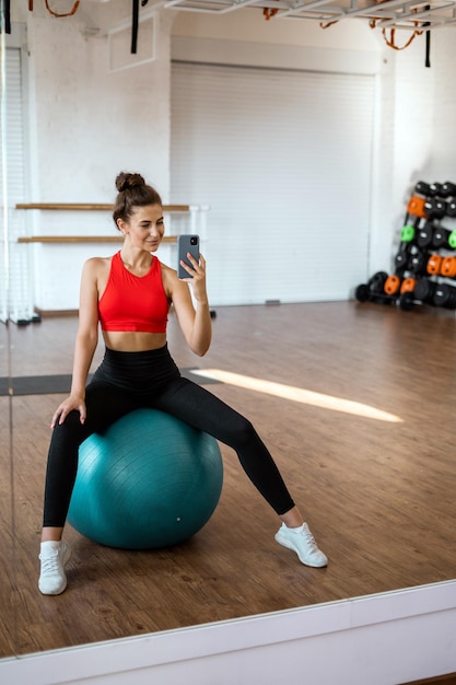 Uma mulher senta-se em uma bola de fitness em uma academia, vestindo um top vermelho e leggings pretas.