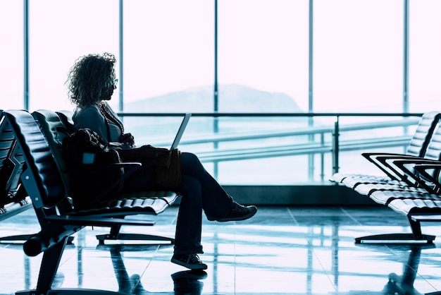 Uma mulher senta-se sozinha no portão do aeroporto esperando o voo para viajar - viagem de negócios ou férias - visão brilhante da janela ao fundo - senhora sentada e esperando atraso avião de partida