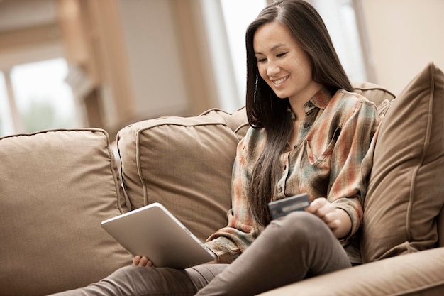 Uma mulher sentada em um sofá em casa segurando um tablet digital e um cartão de crédito comprando online