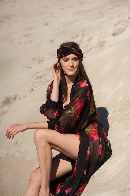 Foto uma mulher sentada em uma duna de areia usando um vestido preto e vermelho com uma bandana.