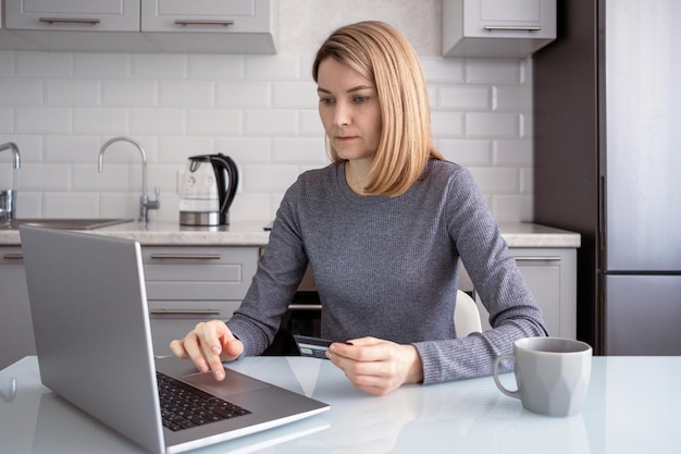 Foto uma mulher sentada em uma mesa na cozinha com um laptop paga as compras no site com um cartão bancário