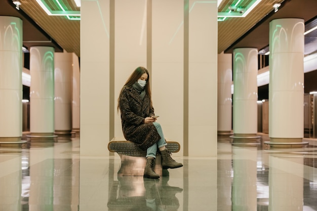 Uma mulher solitária com uma máscara médica para evitar a propagação do coronavírus está sentada usando um smartphone na plataforma do metrô. Uma garota com máscara cirúrgica mantém distância social no metrô.