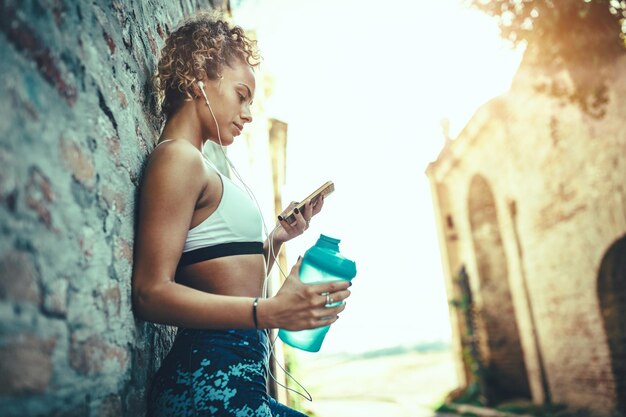 Uma mulher sorridente de fitness jovem com fones de ouvido está fazendo uma pausa depois de correr. Ela está de pé contra a parede e usando telefone inteligente.