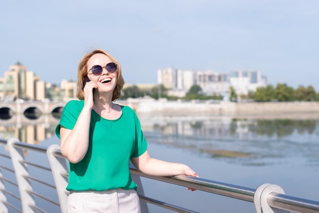 Uma mulher sorridente em óculos de sol falando ao telefone com amigos gesticulando para marcar um encontro na margem do rio na cidade