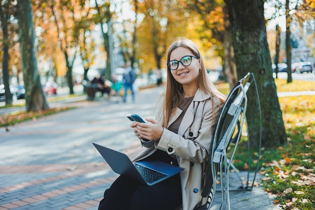 Uma mulher sorridente trabalha com um laptop em um banco no Autumn Park Ela tem um grande sorriso, cabelos longos e grandes olhos azuis Retrato de uma trabalhadora moderna Fundo amarelo do parque