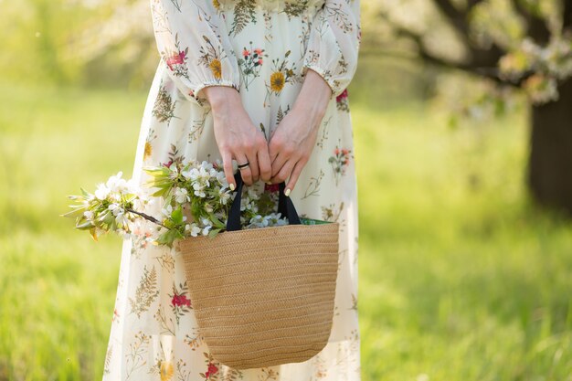 Uma mulher tem nas mãos um saco de vime elegante com flores desabrochando no parque verde. Primavera clima romântico e beuaty da natureza