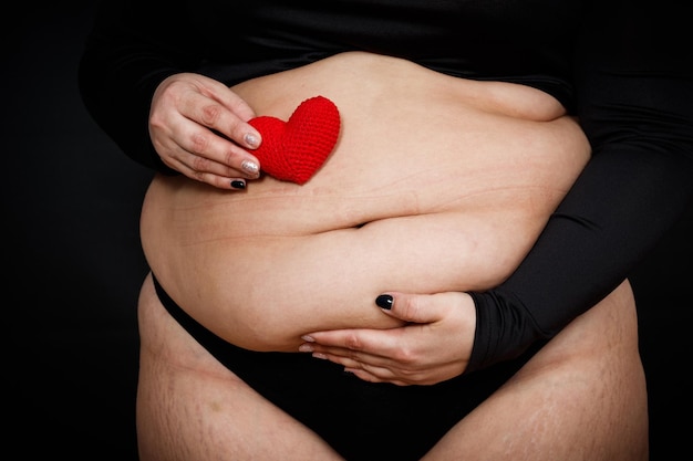 Uma mulher tem um coração vermelho nas mãos em uma barriga gorda em um fundo preto conceito de positividade corporal de pessoa obesa Foto de alta qualidade