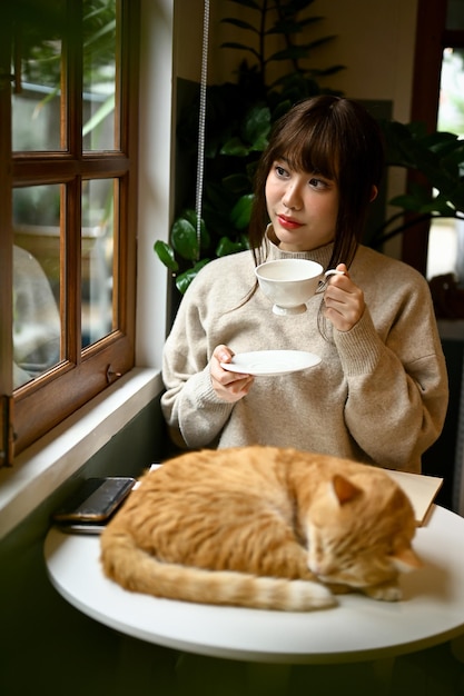 Uma mulher tomando chá em uma mesa perto da janela em sua aconchegante sala de estar com seu gato laranja