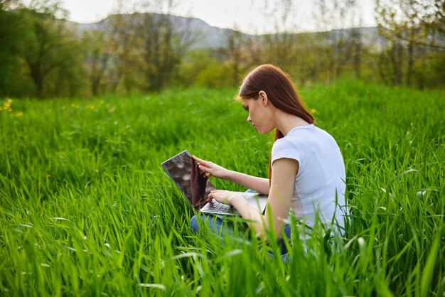 uma mulher trabalha em um laptop enquanto está sentada em alta grama verde na natureza em uma pessoa de um dia de verão ensolarado