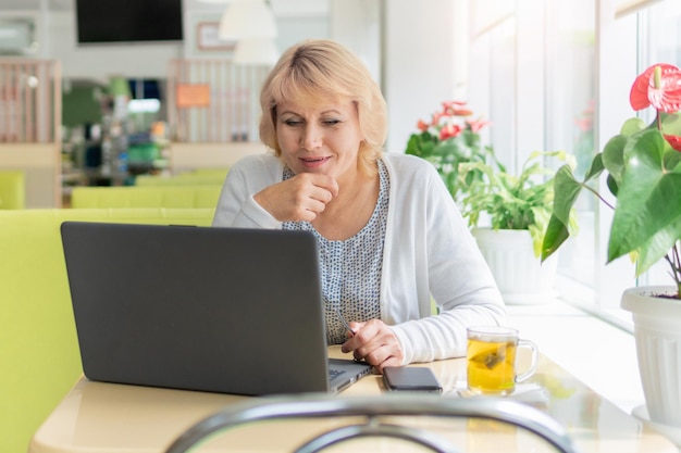 Uma mulher trabalhando com um laptop em uma mesa perto da janela do café Mulher de meia idade freelancer Ela estuda as mídias sociais de e-mail