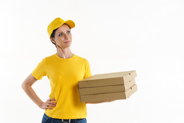 Uma mulher, uma morena, uma entregadora com um uniforme de camiseta e um boné de beisebol amarelo segurando caixas de pizza nas mãos em uma parede branca