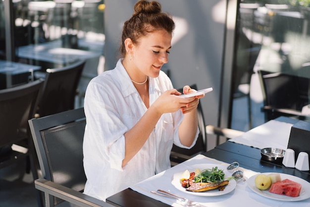 Uma mulher usa um telefone e come almoço ou café da manhã ao ar livre em um café