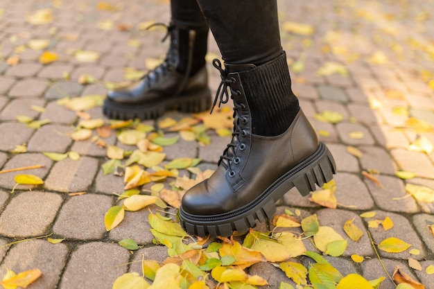 Uma mulher usando botas de couro preto está em um caminho de tijolos com folhas de outono no chão.