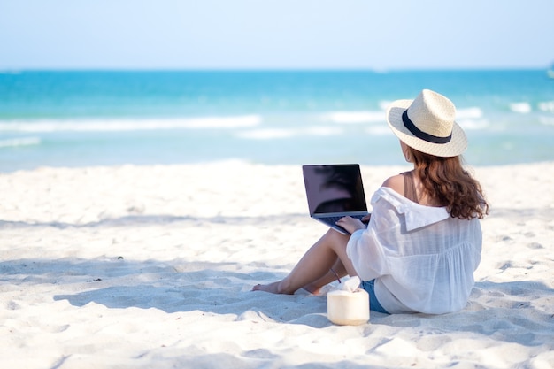 Uma mulher usando e digitando no teclado de um laptop enquanto está sentada em uma bela praia