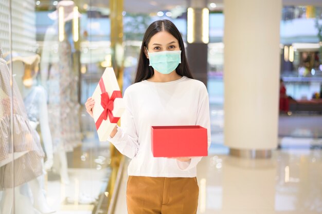 Uma mulher usando máscara protetora segurando uma caixa de presente em um shopping center, sob o conceito de pandemia covid-19, ação de graças e natal.