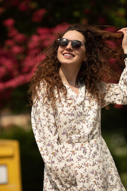 Uma mulher usando óculos escuros e um vestido floral está em frente a uma árvore com flores rosas ao fundo.