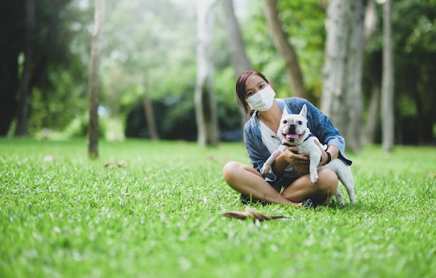 Uma mulher usando uma máscara médica sentada no gramado com um buldogue francês branco