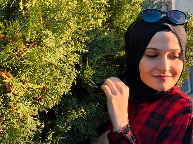 Uma mulher vestindo um hijab e um lenço preto está encostada em uma árvore.