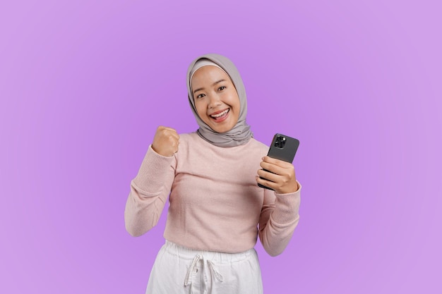 Uma mulher vestindo um hijab está segurando um telefone e sorrindo alegremente depois de receber boas notícias de casa