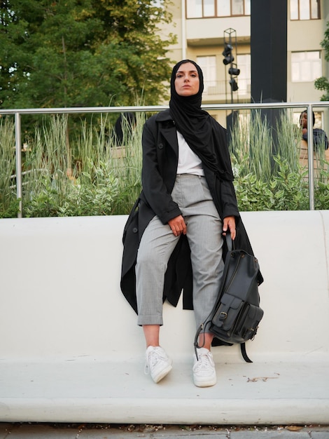 Foto uma mulher vestindo uma jaqueta preta e tênis branco está sentada em uma parede com uma mochila.