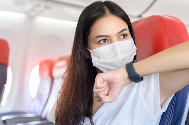 Uma mulher viajando usando máscara protetora a bordo da aeronave usando relógio inteligente, viagem sob a pandemia Covid-19, viagens de segurança, protocolo de distanciamento social, Novo conceito de viagem normal