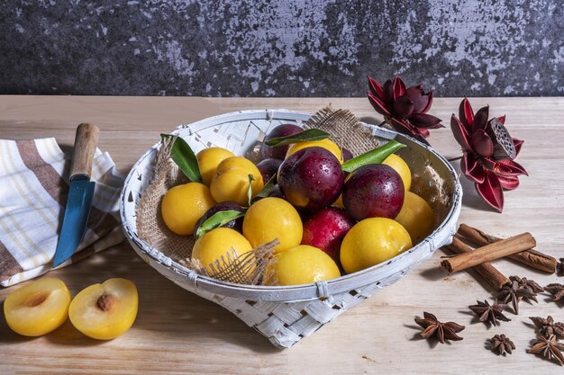 Uma natureza morta com uma cesta cheia de ameixas vermelhas e amarelas maduras junto com pauzinhos de canela e anis doce em uma mesa de madeira leve