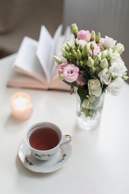 Uma natureza morta de primavera um buquê de rosas em um vaso uma xícara de chá e um livro na mesa branca cartão de saudação de primavera com flores de primavera Fundo romântico