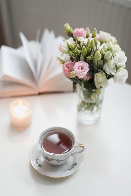 Uma natureza morta de primavera um buquê de rosas em um vaso uma xícara de chá e um livro na mesa branca primavera