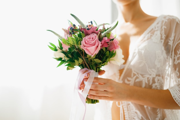 Uma noiva com um penhoar de renda branca segura um buquê de casamento em um quarto de hotel enquanto