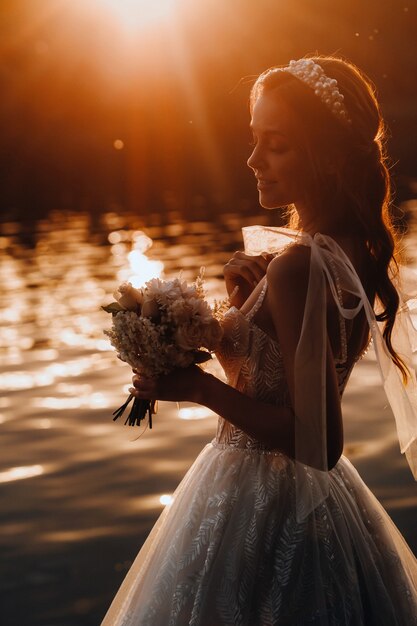 Uma noiva elegante em um vestido branco e luvas está à beira do rio no Parque com um buquê, curtindo a natureza ao pôr do sol. Uma modelo em um vestido de noiva e luvas em um parque natural.