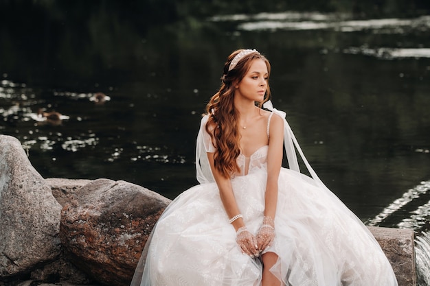 Uma noiva elegante em um vestido branco, luvas e pés descalços está sentada perto de uma cachoeira no parque curtindo a natureza.