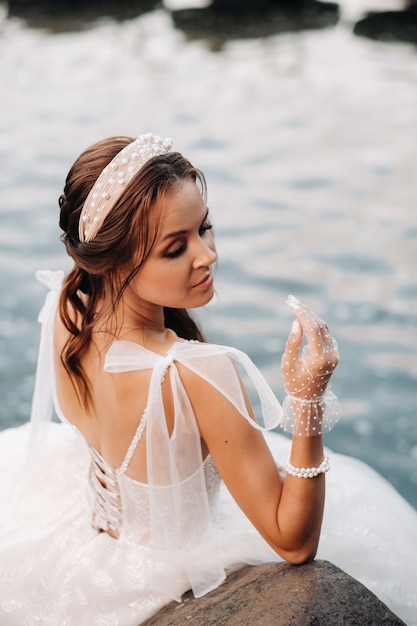 Foto uma noiva elegante em um vestido branco, luvas e pés descalços está sentada perto de uma cachoeira no parque curtindo a natureza.