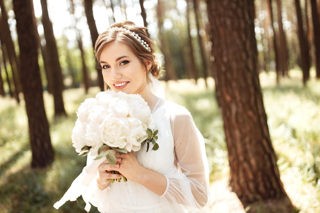 Uma noiva elegante fica na floresta e segura um lindo buquê de casamento com uma peônia nas mãos