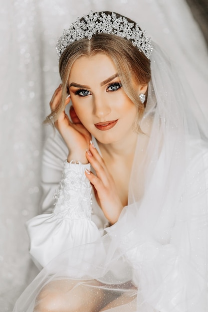 Uma noiva em um terno de seda com um véu posa em seu quarto sentada no chão ao lado de seu vestido de noiva Manhã da noiva Preparação