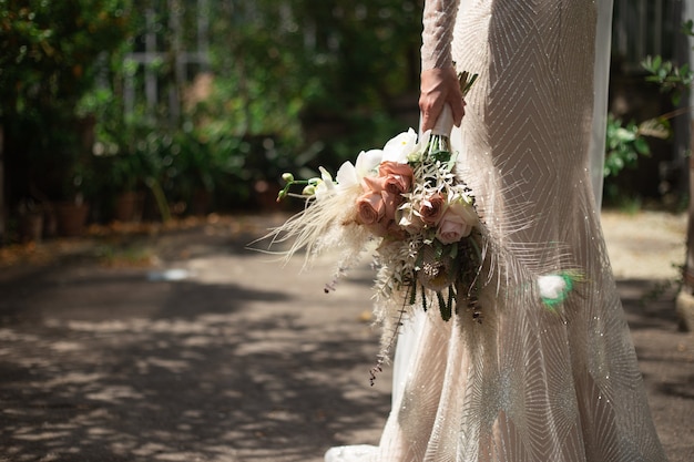 Uma noiva em um vestido de noiva brilhante se levanta e segura um buquê de casamento estilo boho nas mãos.